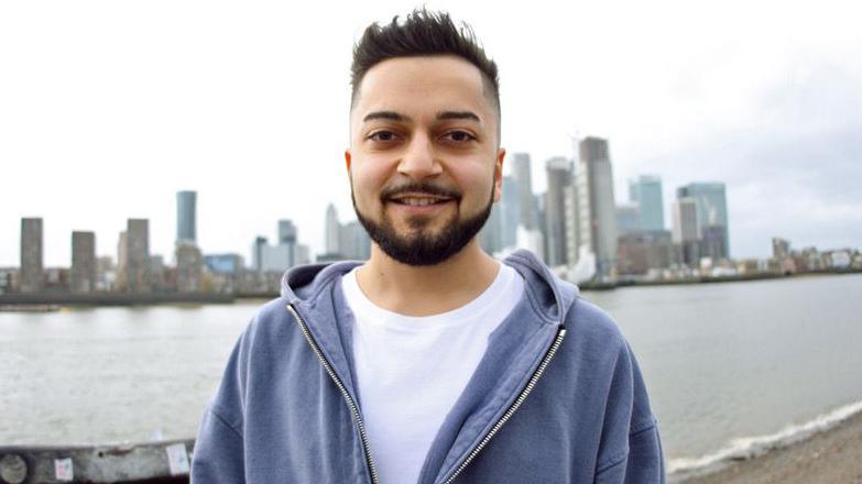 Kishan standing outside wearing a white top and blue hoodie, he is standing with high rise buildings behind him. He is looking straight at the camera and smiling. 