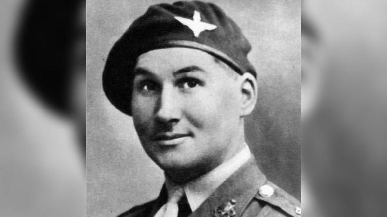 A black and white photo of a male soldier. He is wearing a tie with badges on the collar of his shirt. He is also wearing a hat with a parachute regiment badge on.