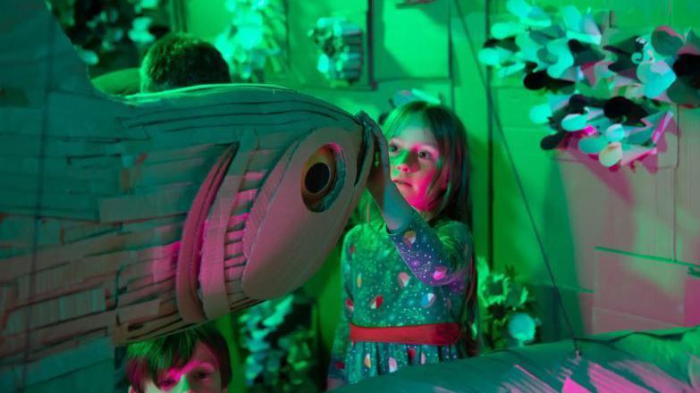 A girl touches the nose of a large fish made from cardboard in a room which is lit in green