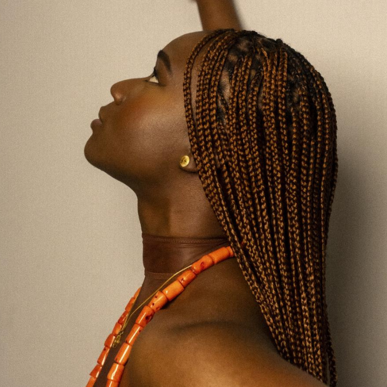 A profiled of a woman with brown hair in braids. She is wearing an orange necklace and gold earring. Her right arm is raised against a plain beige wall.