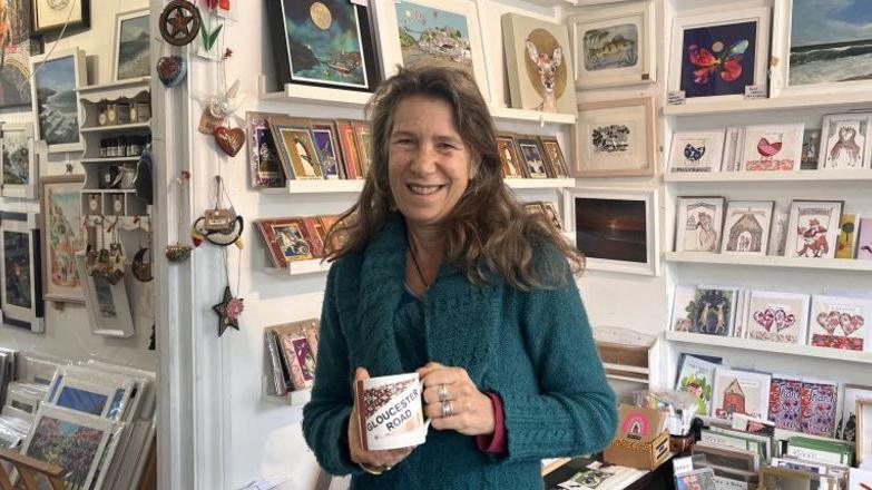 Woman in teal cardigan smiles at camera. She is holding a mug with Gloucester Road on it. She has long brown hair which she is wearing loose and is standing in a shop with lots of art prints on shelves behind her.