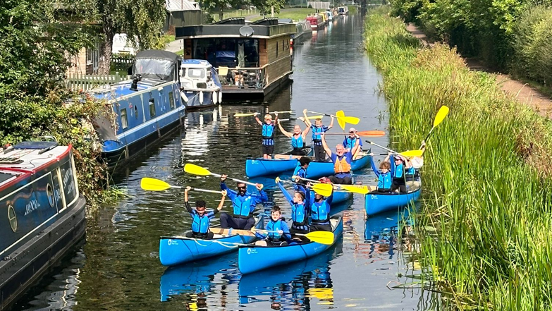 Six blue canoes on a canal with boys raising their paddles over their heads. Narrowboats are moored to the left and reeds are growing in the water on the right of the picture 