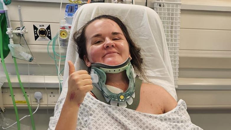 Rebecca O'Malley in hospital wearing a medical gown and a neck brace. She gives the camera a thumbs up and a faint smile.