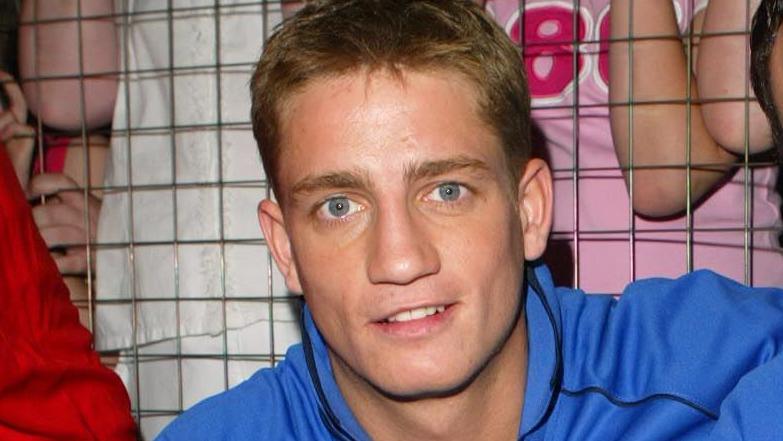 Philip Olivier is pictured during his Brookside years while on SMTV Live. He is wearing a blue hooded jumper with the words "Brookside team". He has short blonde hair and is smiling at the camera.