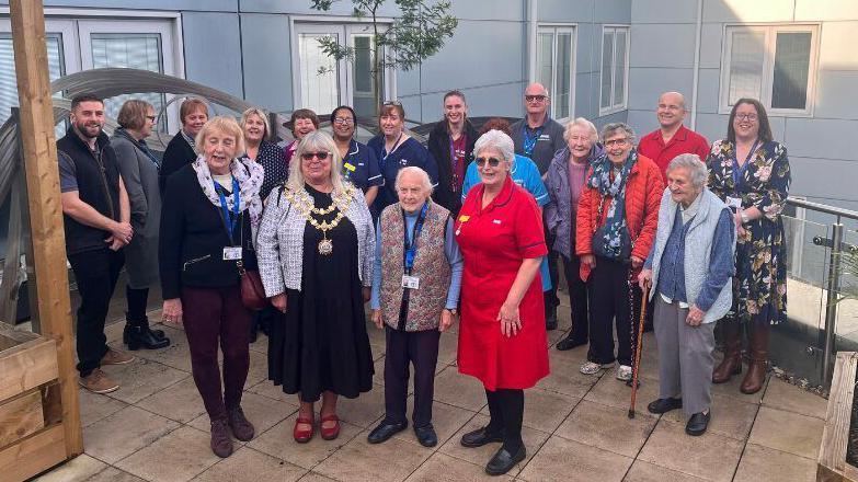 A group of 20 people, including a mayor, hospital staff and volunteers - many of whom are older people - stand on the patio of a balcony garden at a hospital. They are all smiling.