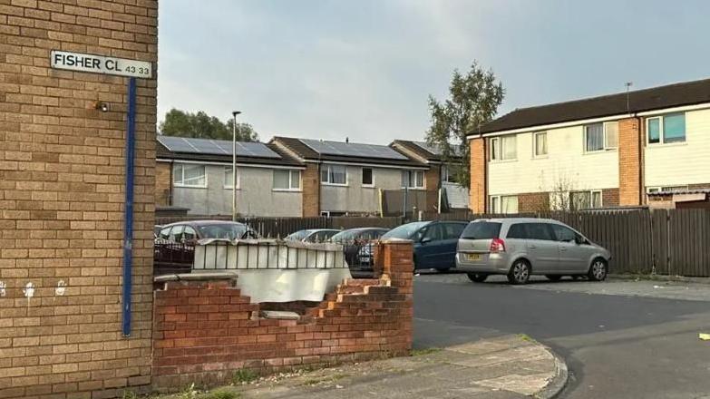 A brick wall with a sign on it reads 'Fisher Cl' which is next to a side street where a number of cars are parked. 