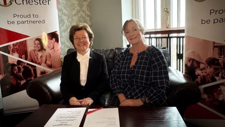 Two women sitting next to each other on a brown sofa with UCM pop up signs in the background. There are two sheets of white paper and a pen on the table in front of them.