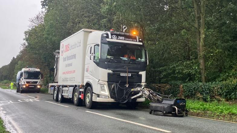 A lorry fitted with the specialist equipment carrying out the work. The lorry is white and it has blasting equipment attached in front of it which is on a road surface