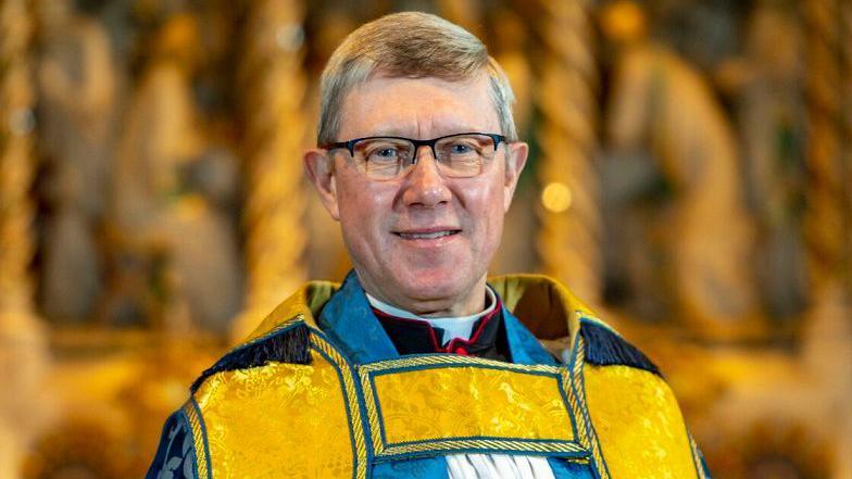 Mark Bonney with short fair hair and glasses wearing yellow vestments