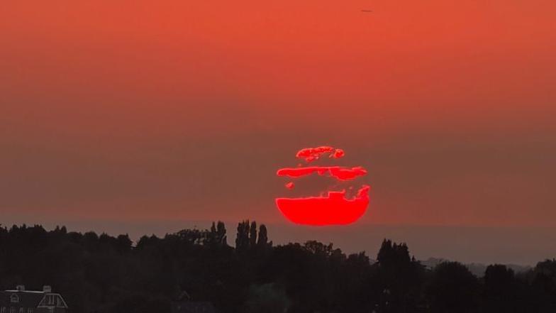 The partially eclipsed Sun appears low above the horizon in Warlingham, Surrey. Dark silhouettes of trees and rooftops sit beneath a red-orange sky, while clouds partially obscure the Sun as it sets.