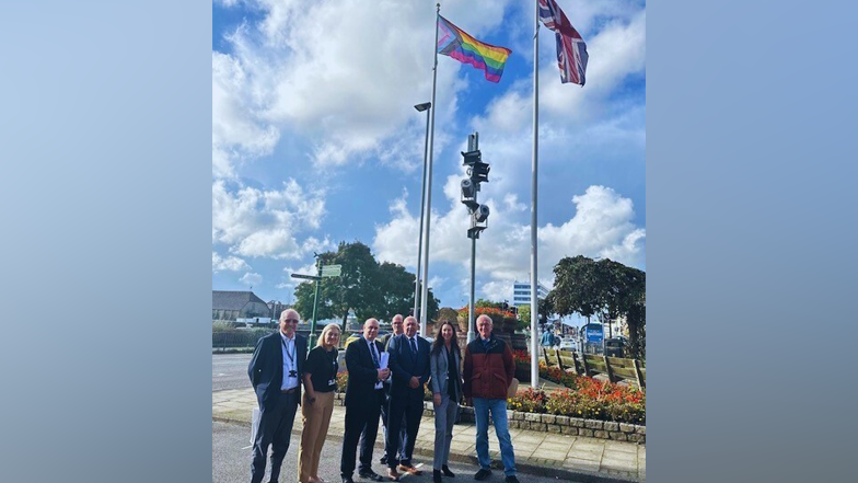 The Progress Pride flag being flown by Great Yarmouth Borough Council. Councillors are stood underneath the flag smiling at the camera. There is another flagpole with a union jack flag flying from it.