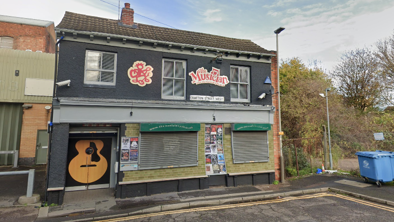 Exterior of the Musician Pub, a two-storey building with a grey exterior and red sign. The front door has an acoustic guitar painted on.