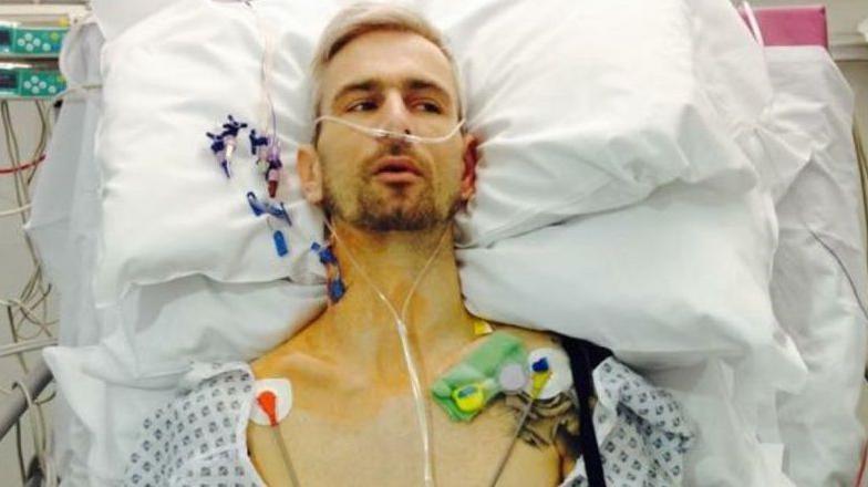 Danny Warren is lying on hospital bed and is wearing a patterned hospital gown. He is surrounded by medical equipment, with various tubes connected to his body.