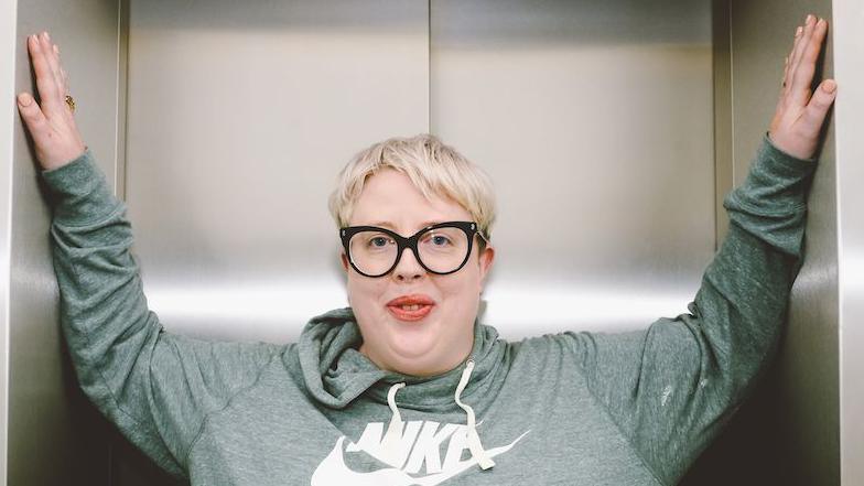 Marea Stamper, aka The Blessed Madonna, poses in a lift at BBC New Broadcasting House, 2022. She is wearing a green hoodie, has short blonde hair and black glasses.