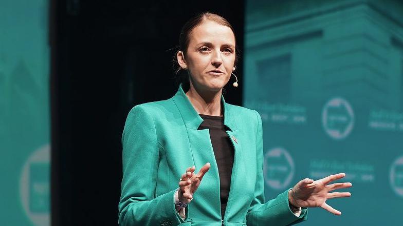 Maria Bowtell dressed in a turquoise suit, matching the colour of the Reform UK backdrop, addresses a party conference. Her hair is scraped back and she is making a point with her hands.