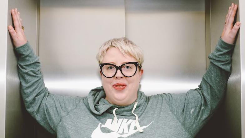 Marea Stamper, aka The Blessed Madonna, poses in a lift at BBC New Broadcasting House, 2022. She is wearing a green hoodie, has short blonde hair and black glasses.