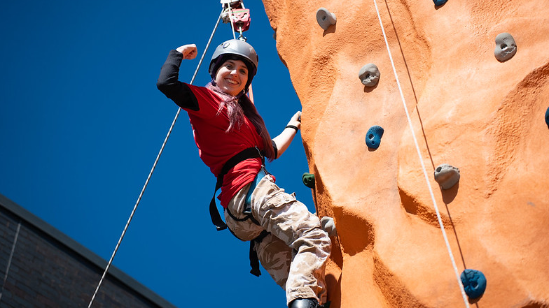 Ava is at the top of an orange climbing wall, wearing a harness and helmet and smiling to camera with her fist in the air in triumph. There is a beautiful blue sky in the background