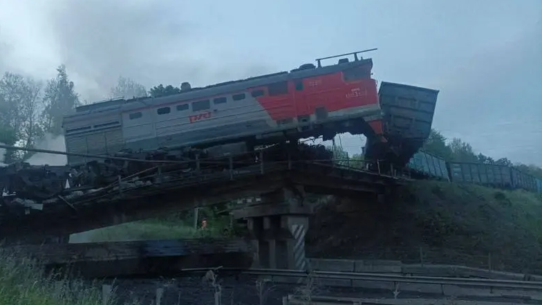 A freight locomotive is flipped upside down after a rail bridge collapsed. the picture shows a mangled train and a burnt out bridge