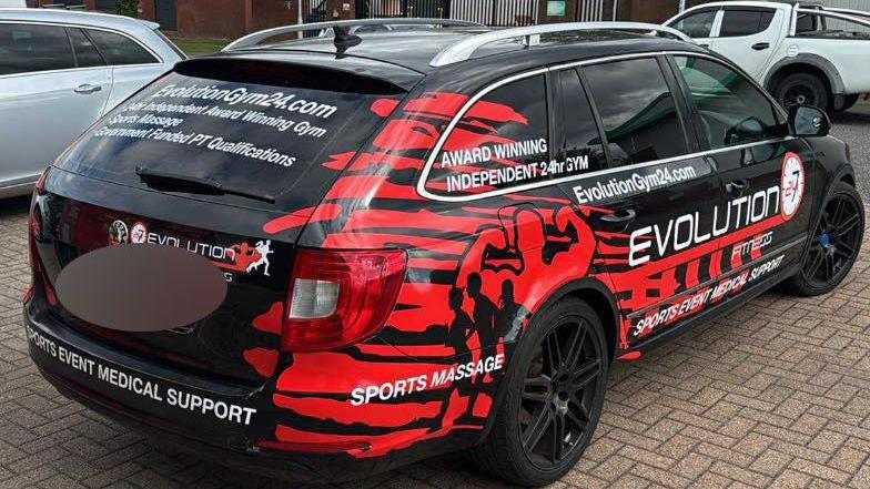 The four-door car is black with red decal displaying information about the Evolution Fitness gym, including an email address and website url.