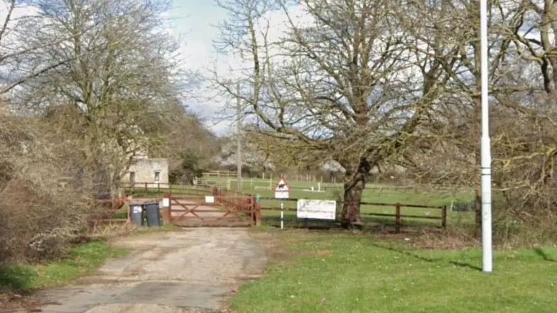 A driveway leads up to a wooden gate before a stone outbuilding. There are a number of fields to the gate's right. On the fields are two black wheelie bins, a sign and a number of trees. 