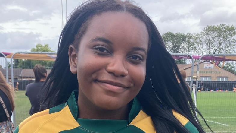 Anisha has long black hair and is wearing a green and yellow striped sports top. She is smiling and standing in front of the rugby pitch.