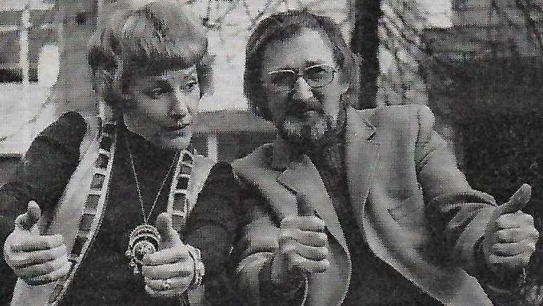 A black-and-white photo of a woman and a man sitting next to each other with their thumbs up. The man has long hair and a beard and wears a jacket and glasses. He is looking straight ahead. The woman has short hair and wears a dark top, sleeveless jacket and large medallion. She is looking towards the man.