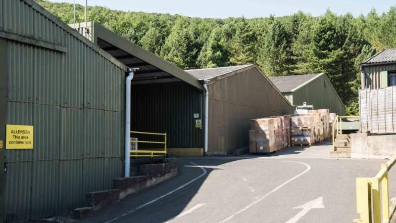 A section of the former Yeo Valley Farm site in Cheddar. It shows an entrance driveway turning left into a corrugated metal shed building. There are also wrapped crates of dairy products. The site is bordered by a forest of mature trees stretching uphill. 