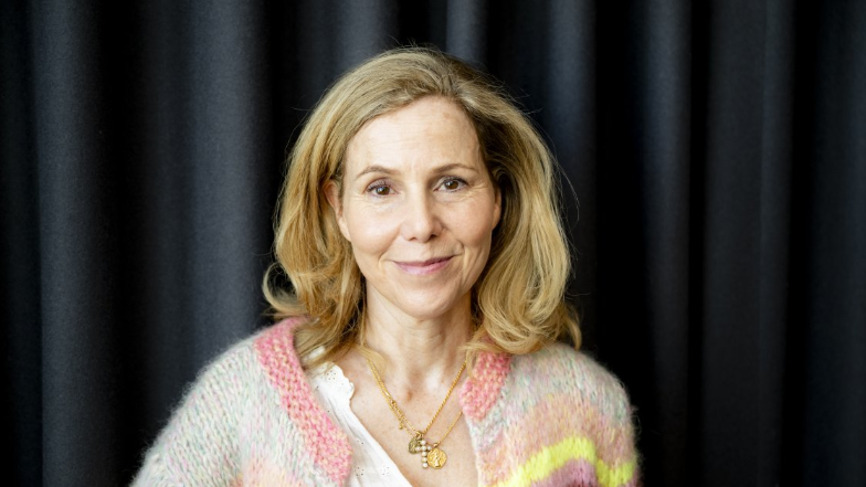 Actress Sally Phillips smiles at the camera in front of a black curtain. She has shoulder-length blonde hair, brown eyes, and wears a multi-colour pastel cardigan.