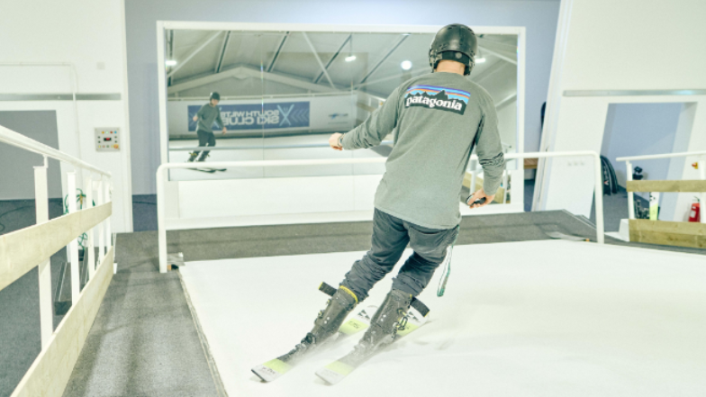 Someone using the ski slope at the site. They are wearing a brown top and ski trousers and can see themselves in a large mirror screen