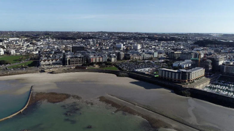 Seen from above is the St Helier waterfront with the sea in the foreground, a beach in the middle ground and buildings and sky in the background. 