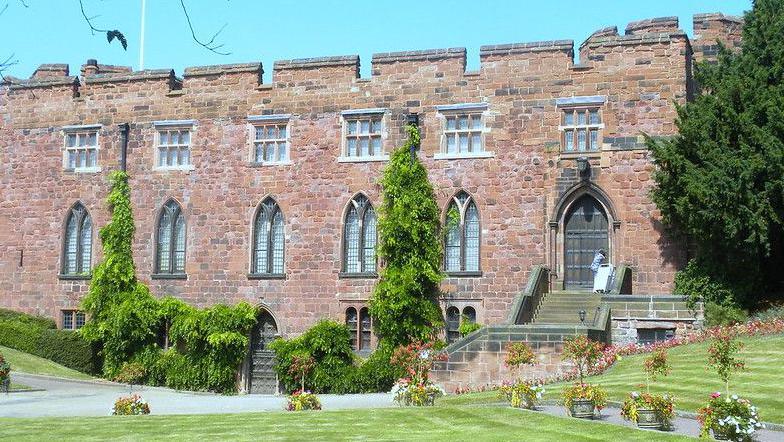 A grand old building made of red stone, with well-trimmed lawns to the front and sides