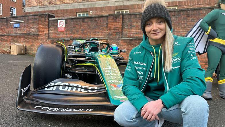 A blonde woman in a bobble hat, jeans and green jacket, crouched in front of an F1 replica car which has Aramco written on it.