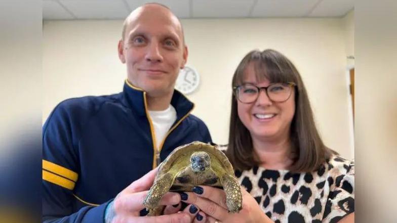 A man with a bald head and blue jacket, and a women with brown hair and glasses, hold up a tortoise. They are smiling.