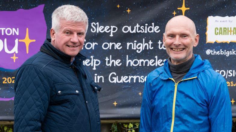 Wearing trousers and raincoats and smiling, David Stoddart and Graham Merfield stand side by side in front of a banner which advertises the Skipton Sleep Out. 