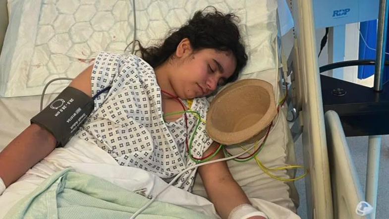 Maya, who is lying on a hospital bed and is wearing a white, patterned hospital gown, is attached to medical equipment. There is a sick bowl next to her. 