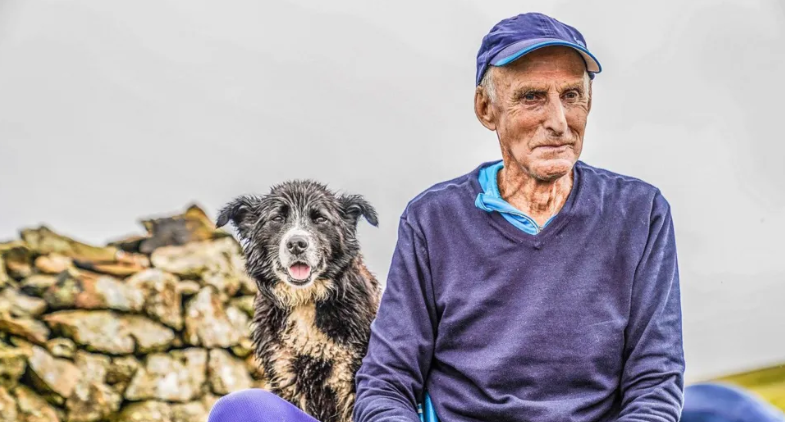 A man with white hair and a blue cap sitting next to a wet sheepdog and pile of stones. He is looking to the side wearing a blue jumper. The dog is looking into the camera.