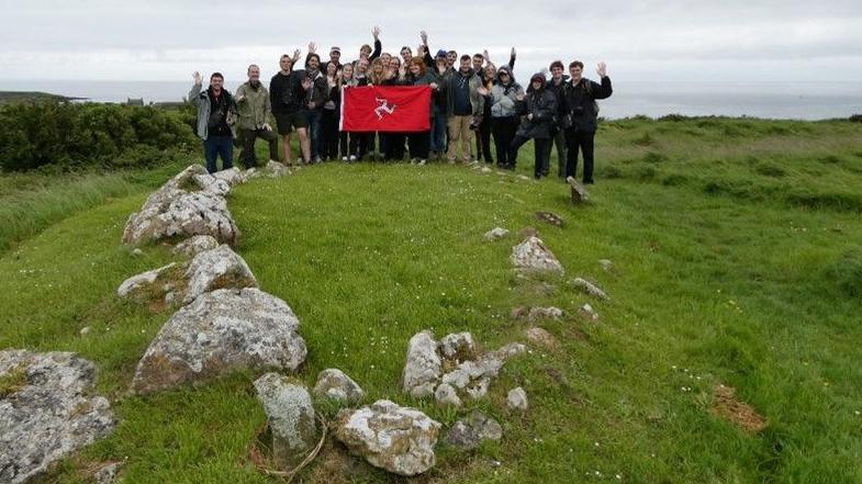 A group of students in a green hillside holding up a Manx flag. They are standing in front of stones marking the outline of a Viking boat burial.