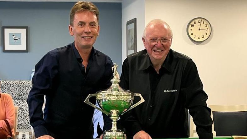 Ken Doherty, left, and Dennis Taylor, right, standing in front of a snooker table with a replica championship trophy on it. They are both looking into the camera, smiling, and wearing dark long sleeve clothing. 