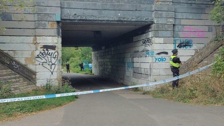 Melrosegate underpass with a police cordon in front of it and a police officer wearing a hi-vis jacket and police hat, looking away from the camera. There is graffiti on the bridge of the underpass and other officers are seen in the distance behind the bridge.