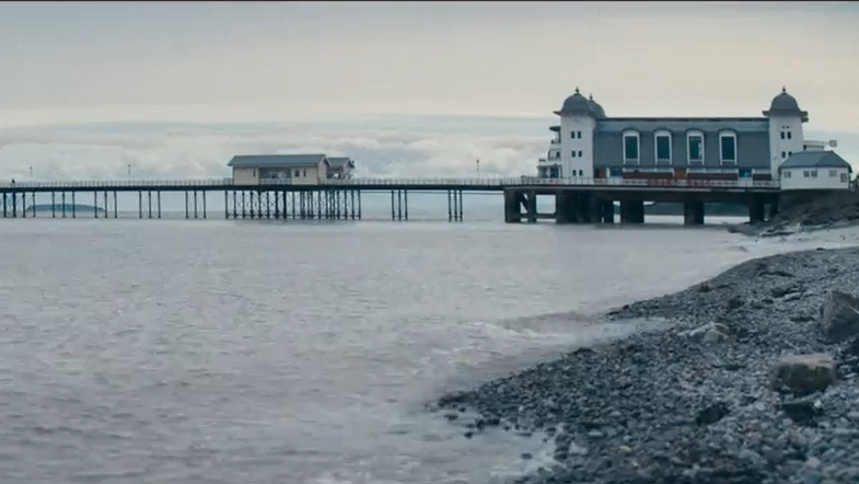 A cloudy view of Penarth Pier. The tide can be seen coming in high on the grey rocks. A white and grey building is seen at the entrance of the pier. 