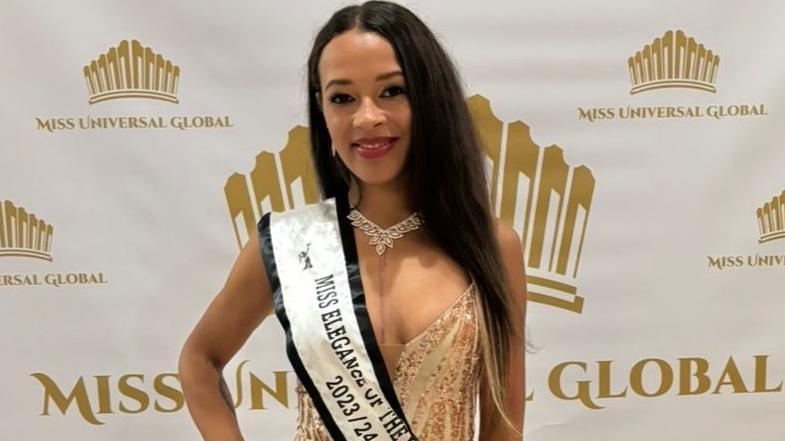 Crystal Priscott. She has long dark hair and is wearing red lipstick. She is in a beige sparkly dress and is wearing a white sash over her shoulder with "Miss Elegance of the World 2023/24" written across it. The scar on her chest from open heart surgery can be seen. She is posing with her hand on her hip in front of a background with the pageant's branding on it.