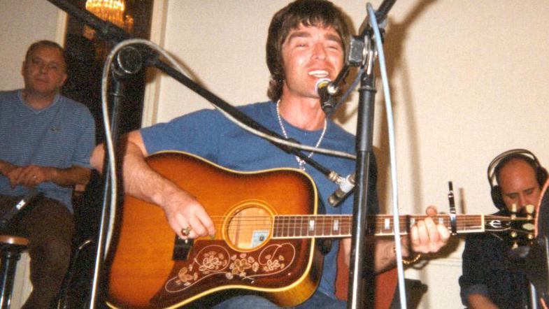 Noel Gallagher sat on a chair singing and playing a sunburst Epiphone acoustic guitar with two microphones set up in front of him. His hair is down the back of his neck.