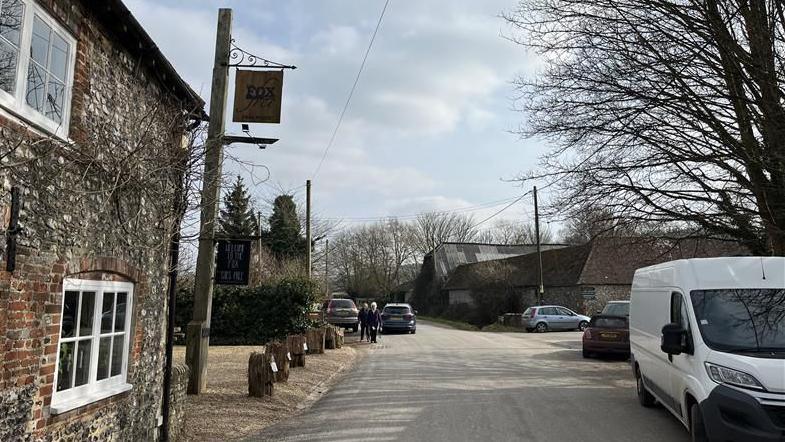 The Fox Goes Free pub is a flint and brick walled pub with small windows and low door frames. It is next to the main narrow lane through the village which has cars parked on it. And there is a barn on the other side of the road