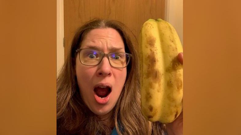 Kate Wright wearing glasses with brown hair looking shocked holding the "double" banana. It looks like the skin has not separated from two bananas.