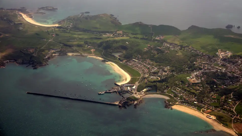 An aerial photograph of Alderney. Green land is visible with a beach and blue water surrounding it.