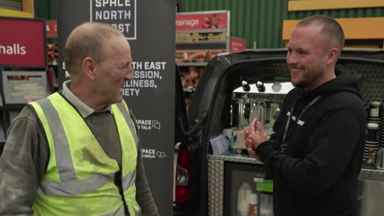 James Fildes, in black hoody and trousers is smiling and talking to a man in khaki jumper, high visibility vest and jeans. They are standing in front of the van in Durham, which is equipped with a coffee machine, fridge with milk and cream and disposable paper cups.