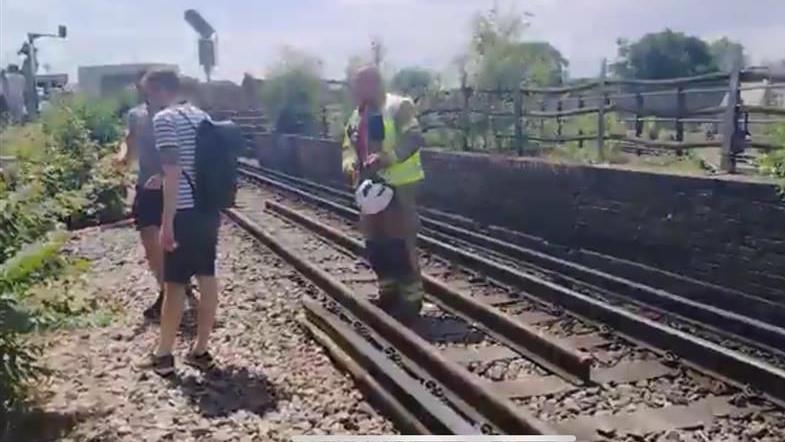 Two train passengers walk alongside train tracks as firefighter guides their direction.