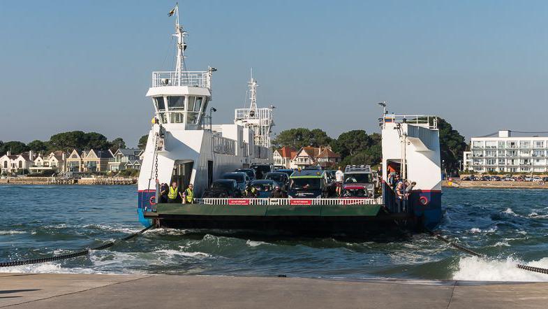 The Sandbanks Ferry, loaded with cars, arrives at Studland. Houses on Sandbanks across the water are in the background.