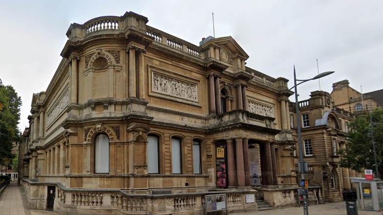 Wolverhampton Art gallery is a brown-beige stone building with people carved into a design near the roof. There are pillars around the entrance which has a small flight of steps. There is also a balcony above the entrance which also has brown pillars on it.