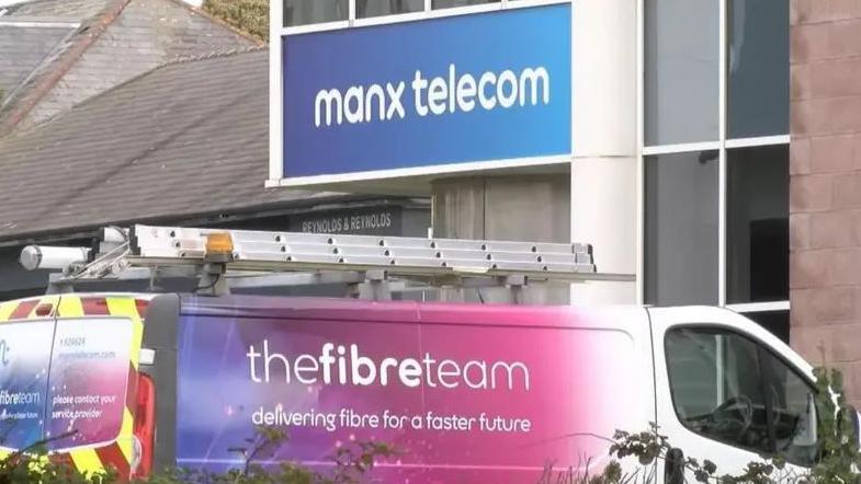 A van with colourful Manx Telecom branding on its rear and driver side panel and a ladder on its roof. Its parked outside a building with blue and white sign that reads Manx telecom.